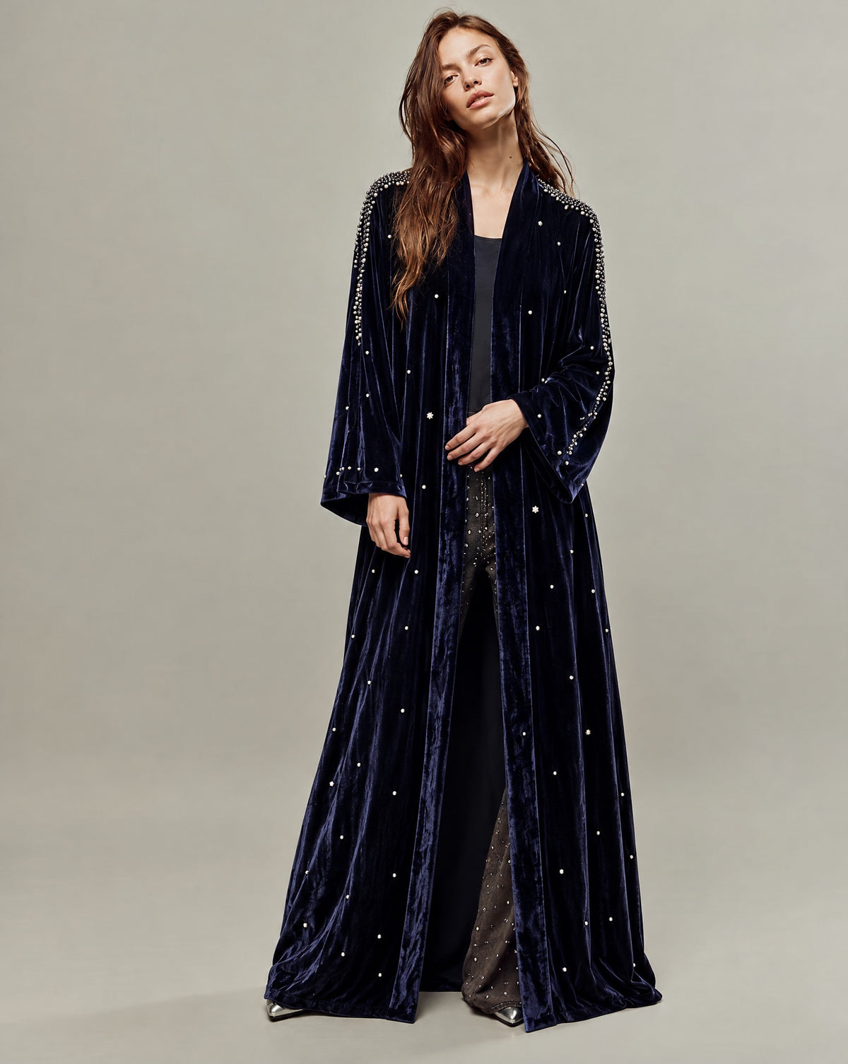 Model with long hair in an open navy velvet abaya studded with crystals on the shoulders, sleeves and body, over a black top and sparkly trousers, on a plain grey studio background