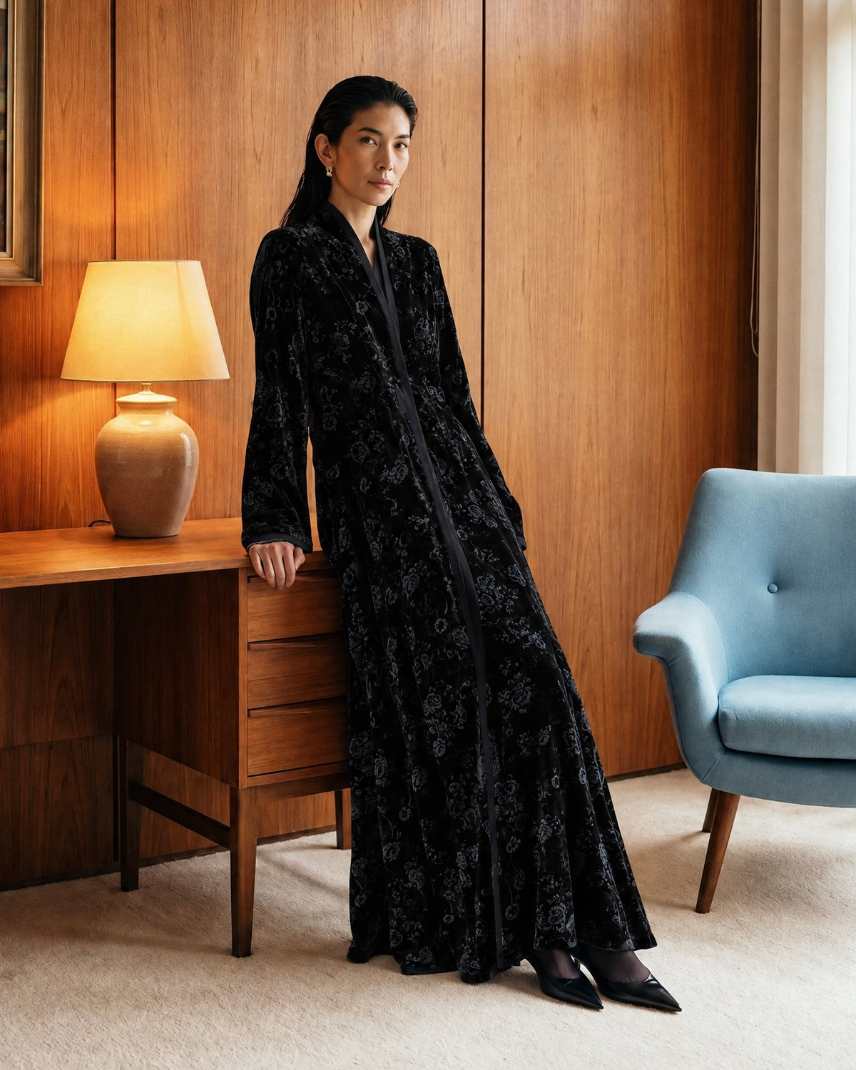 Model in a long black floral velvet abaya with black pointed heels and small gold earrings, leaning on a wooden desk with a lamp, in a wood-panelled room with a landscape painting and a pale blue armchair