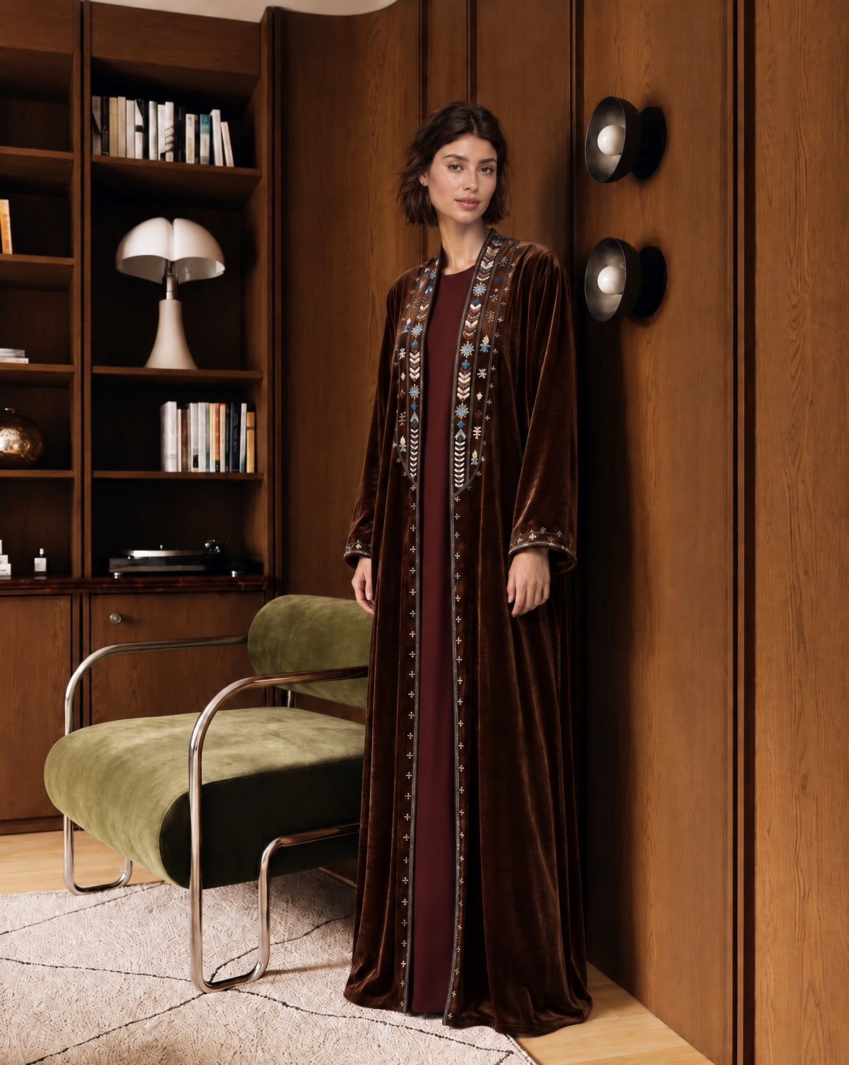 Model with short dark hair in an open brown velvet abaya with multicolour geometric embroidery on the lapels and cuffs over a burgundy dress, standing beside an olive velvet armchair in a wood-panelled room with bookshelves