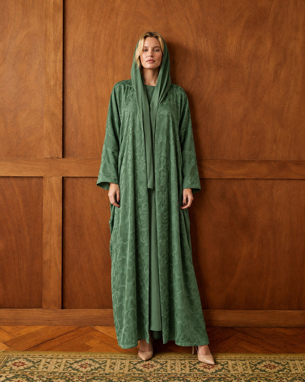Model in a green washed-silk hooded abaya standing against a wood-panelled wall