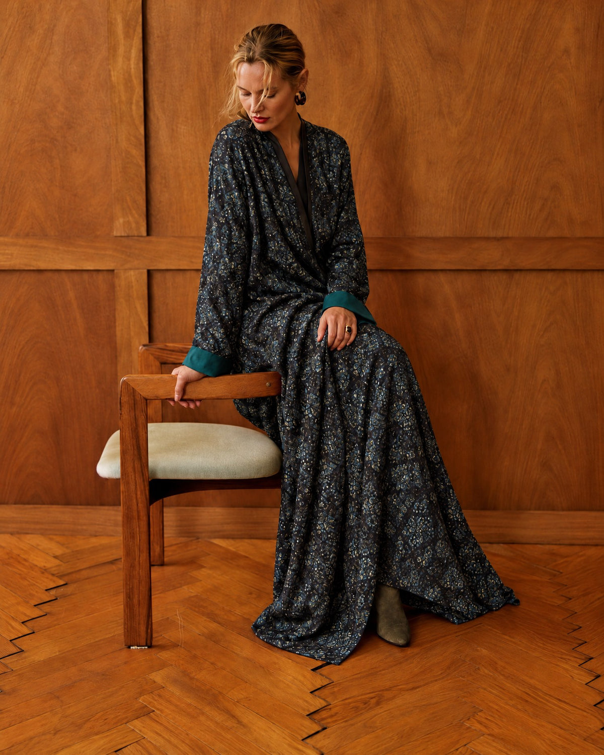 Model in a dark printed jacquard abaya with teal cuffs, seated on a wooden chair in a wood-panelled room