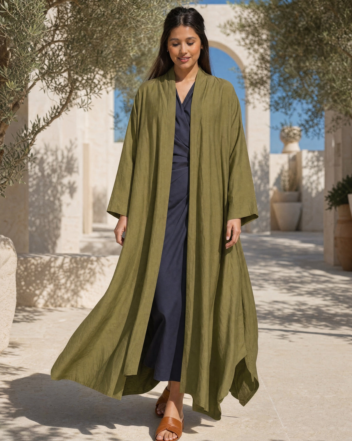 Orange Blossom Sylvia olive linen abaya worn open over a navy dress, walking under olive trees