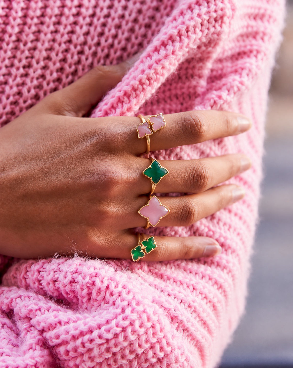A hand wearing four gold rings with pink and green clover-shaped stones, on a pink knit sleeve