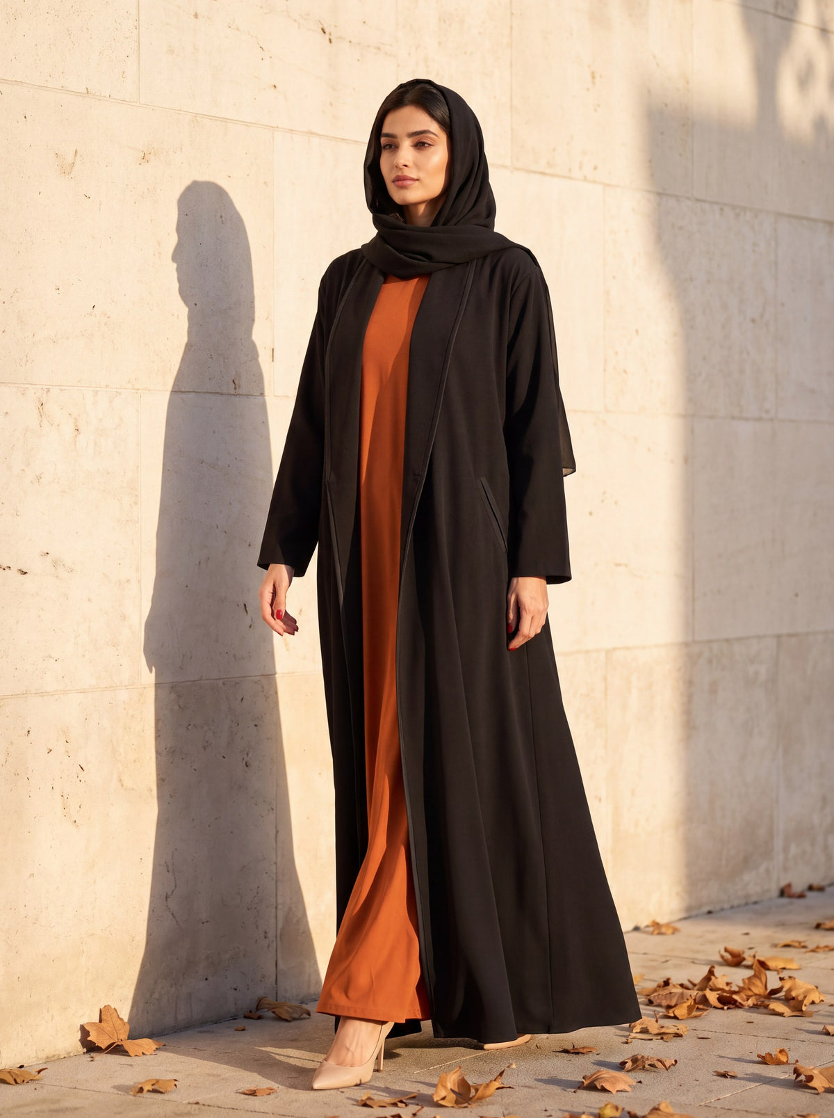 Black crepe abaya coat over a burnt-orange dress, by a sunlit limestone wall