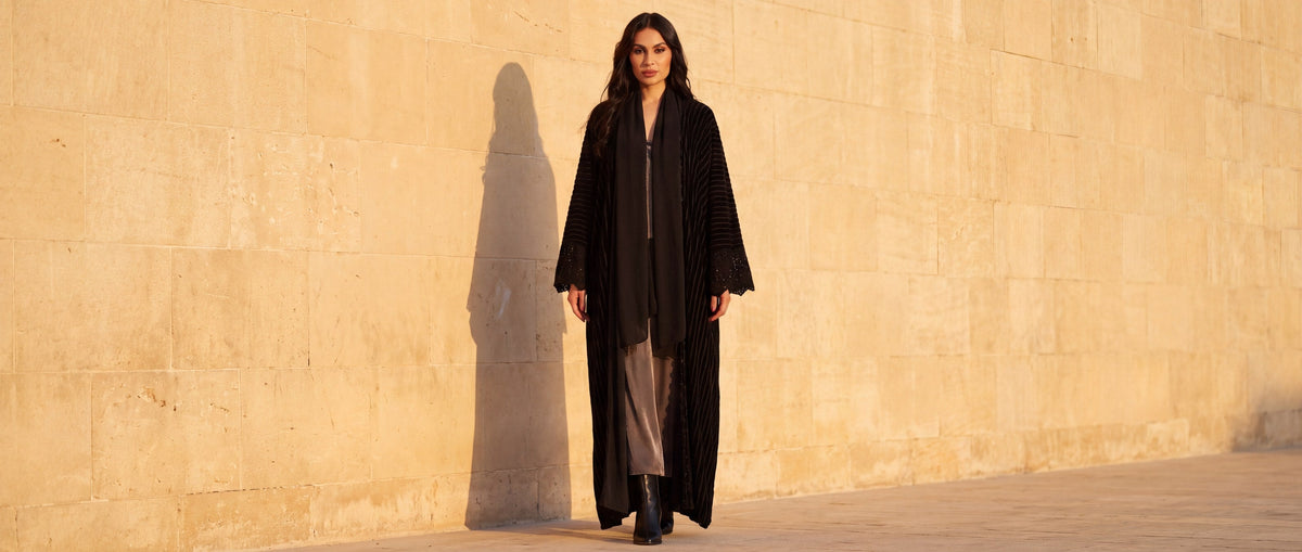 Model in the Onyx black striped-velvet abaya with crochet cuffs and a black stole, walking along a sunlit limestone wall