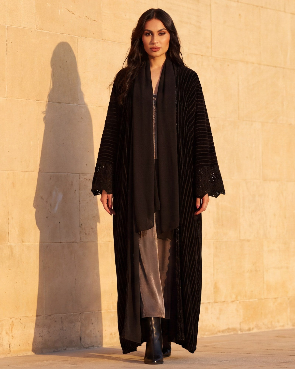 Model in the Onyx black striped-velvet abaya with crochet cuffs and a black stole, walking along a sunlit limestone wall