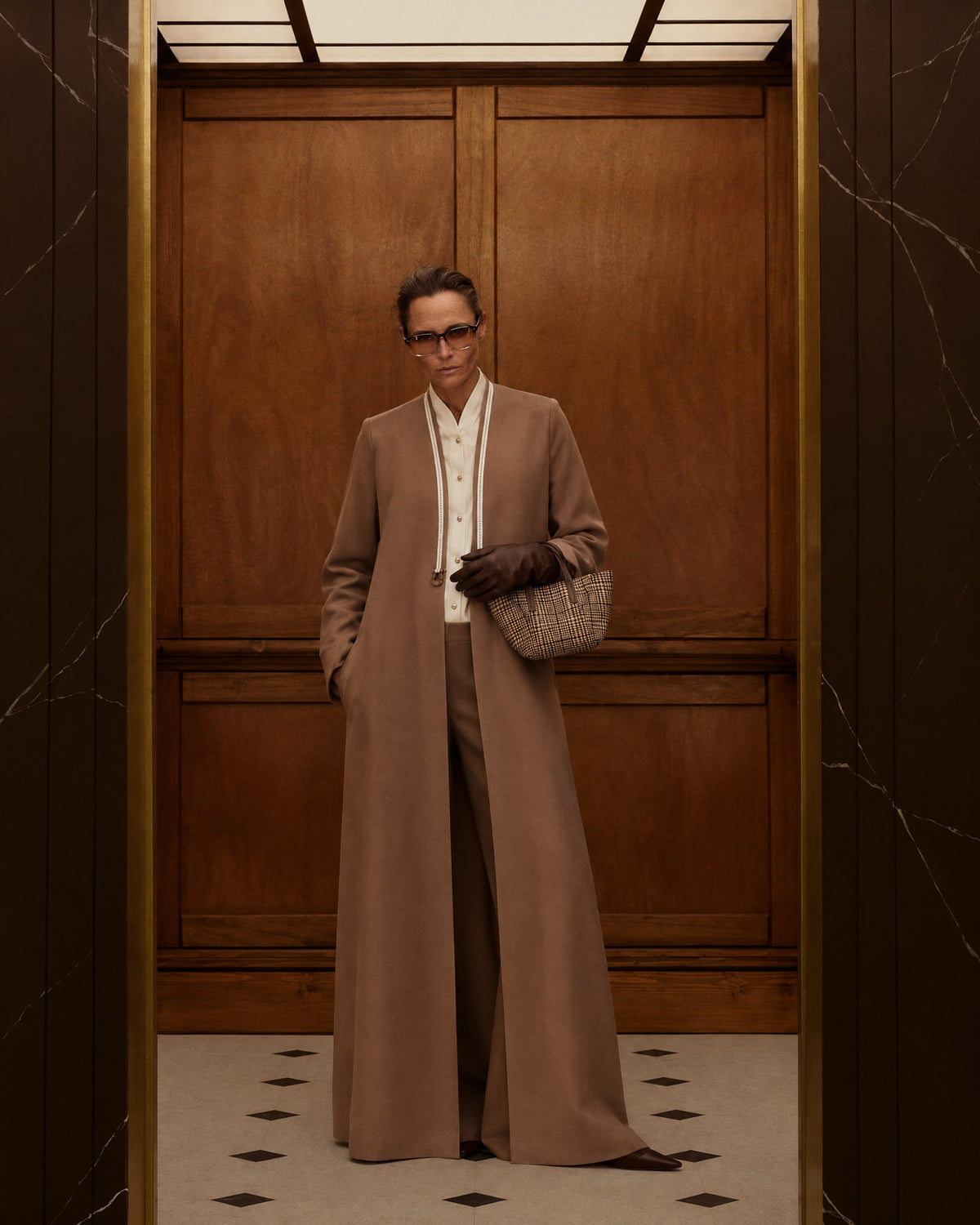 Model in the CODE-0425 tan viscose abaya coat with cream piping, glasses and brown gloves, in a wood-panelled elevator