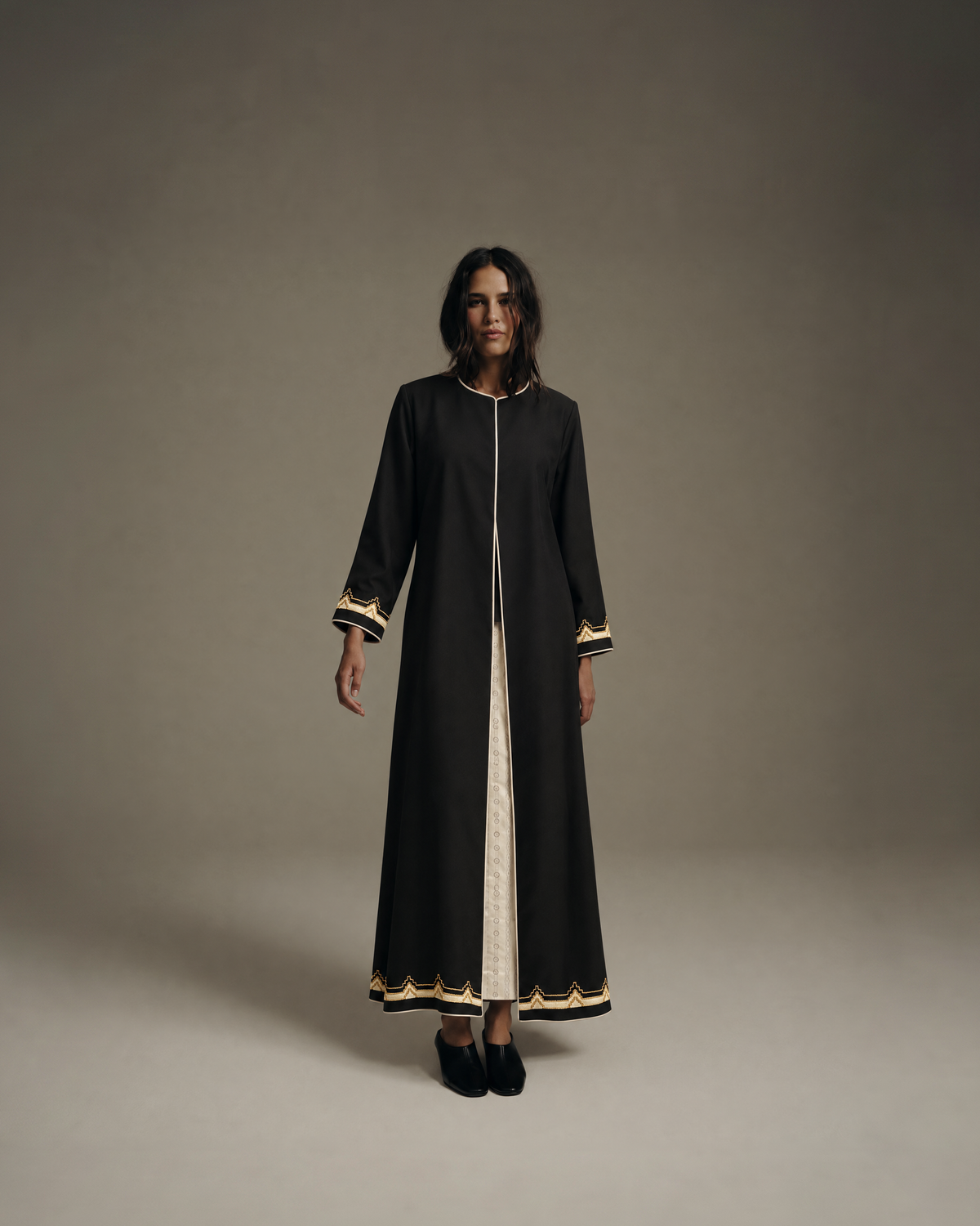 Model with shoulder-length dark hair in an open black abaya with cream piping and gold and cream scalloped trim at the cuffs and hem, over a long cream dress, with black mules, standing full length on a plain grey studio background