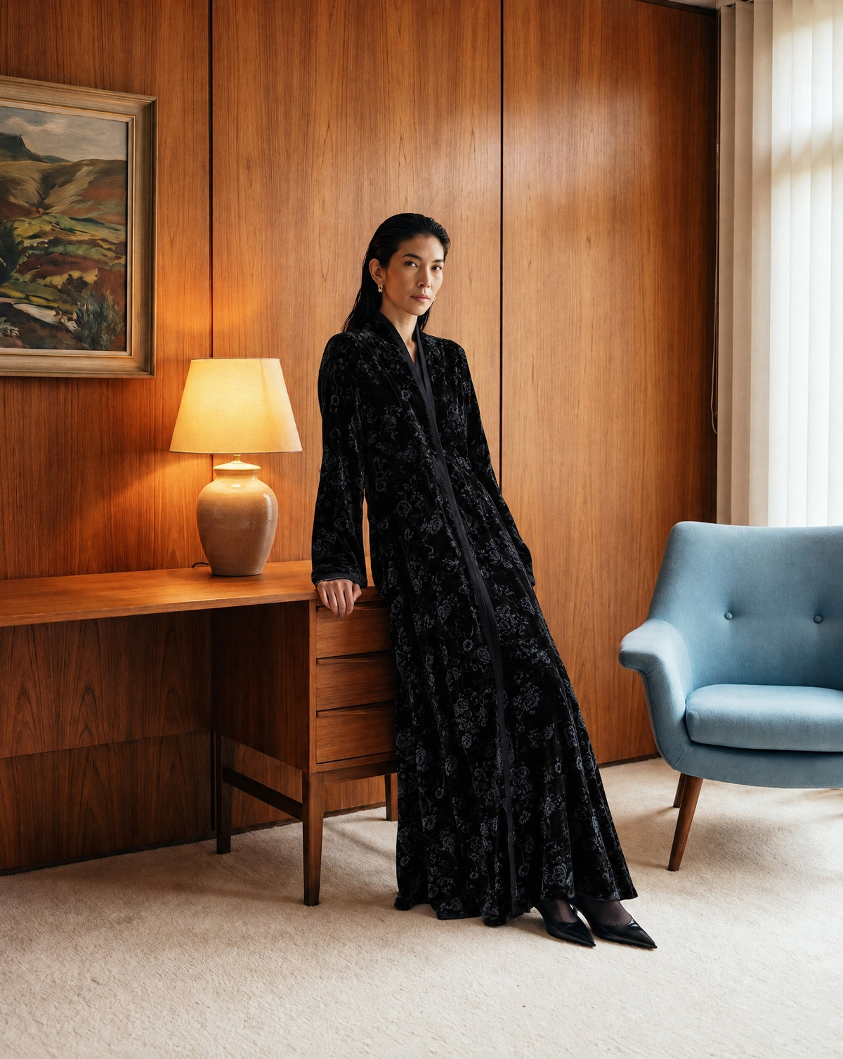 Model in a long black floral velvet abaya with black pointed heels and small gold earrings, leaning on a wooden desk with a lamp, in a wood-panelled room with a landscape painting and a pale blue armchair