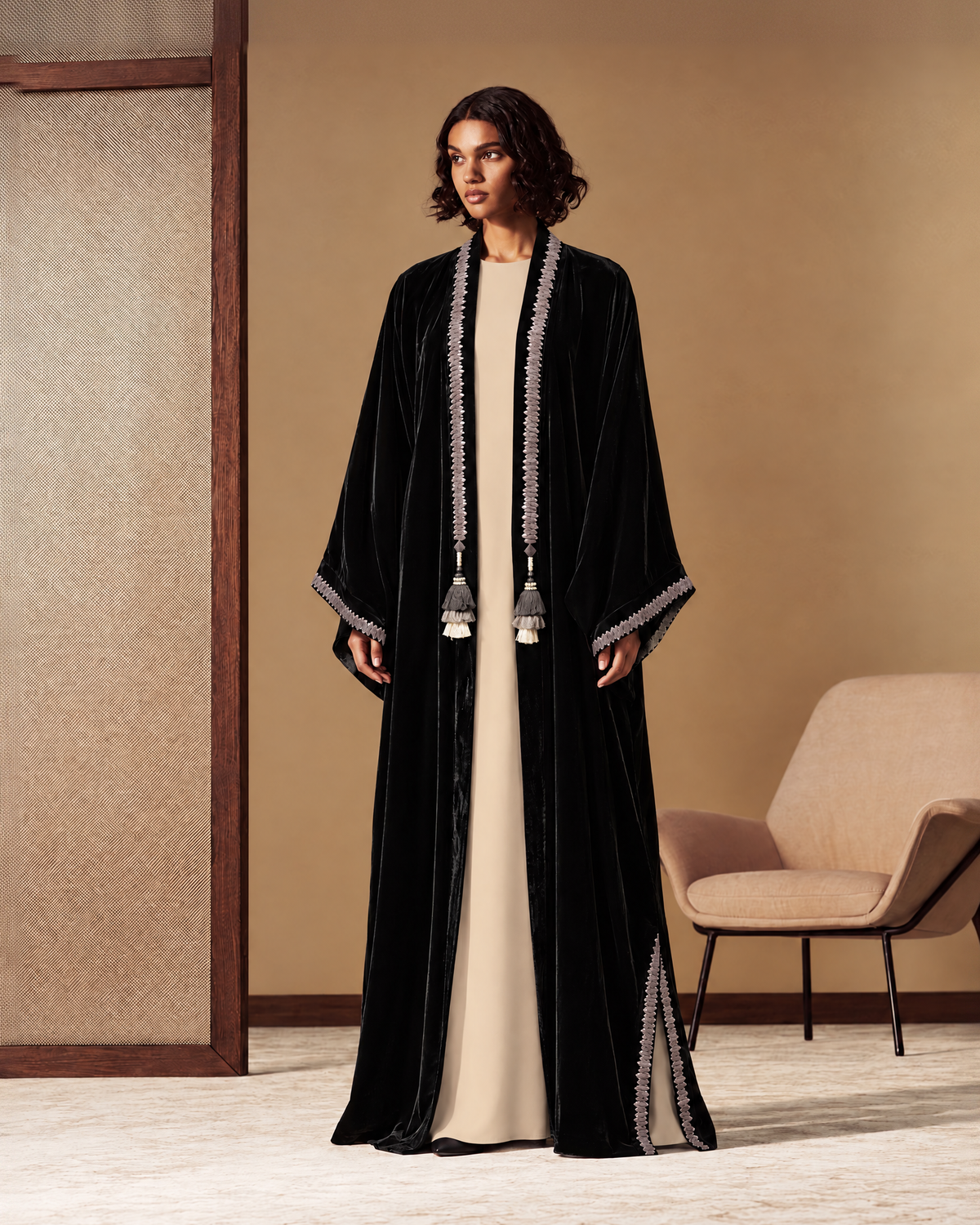 Model with shoulder-length dark hair in an open black abaya with cream piping and gold and cream scalloped trim at the cuffs and hem, over a long cream dress, with black mules, standing full length on a plain grey studio background
