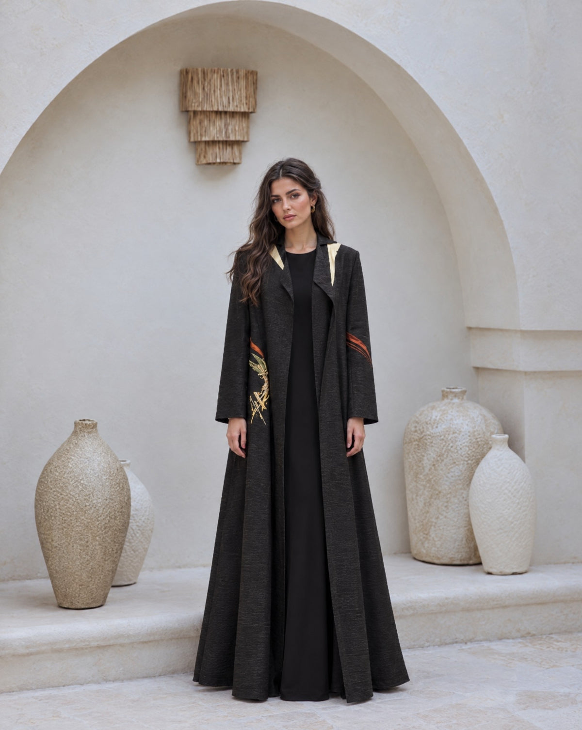 Model in a black jacquard coat abaya with cream lapel inserts and gold embroidery, in a white courtyard