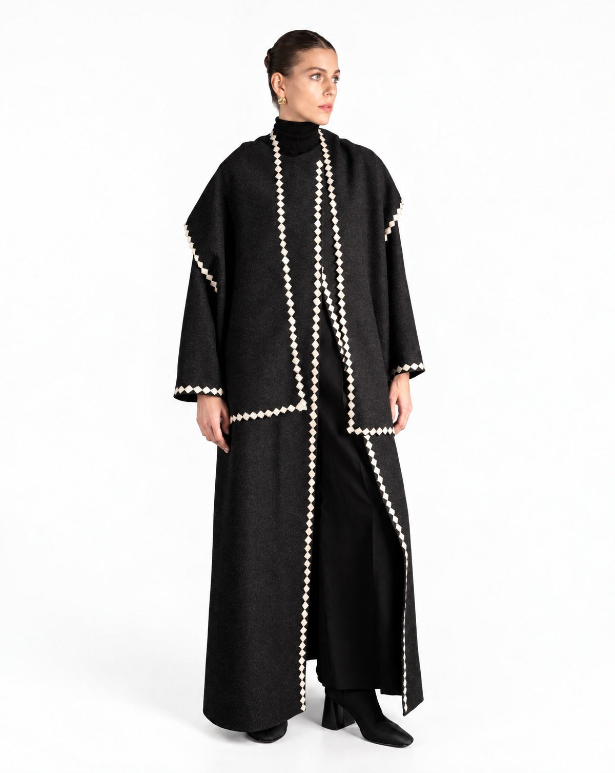 Model in a long black wool coat abaya with cream scalloped trim, worn open over black, against a white wall