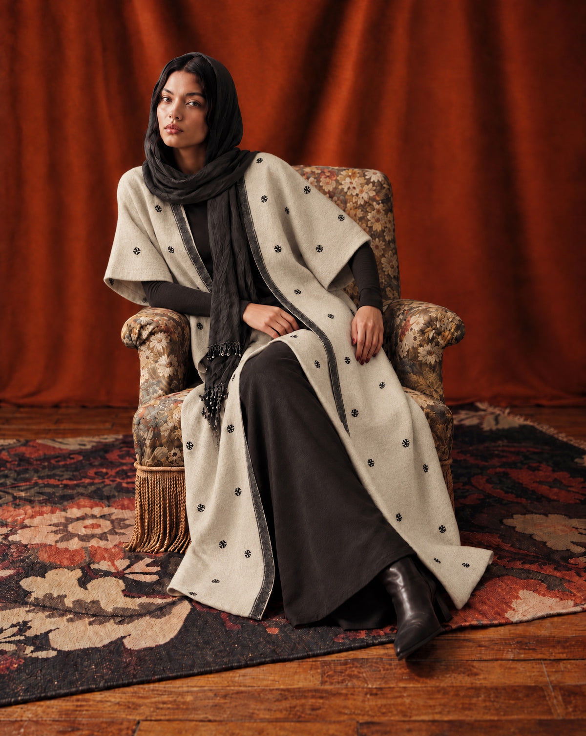 Model seated in a floral armchair in a cream wool abaya coat with black flower motifs and a black border, against a rust curtain