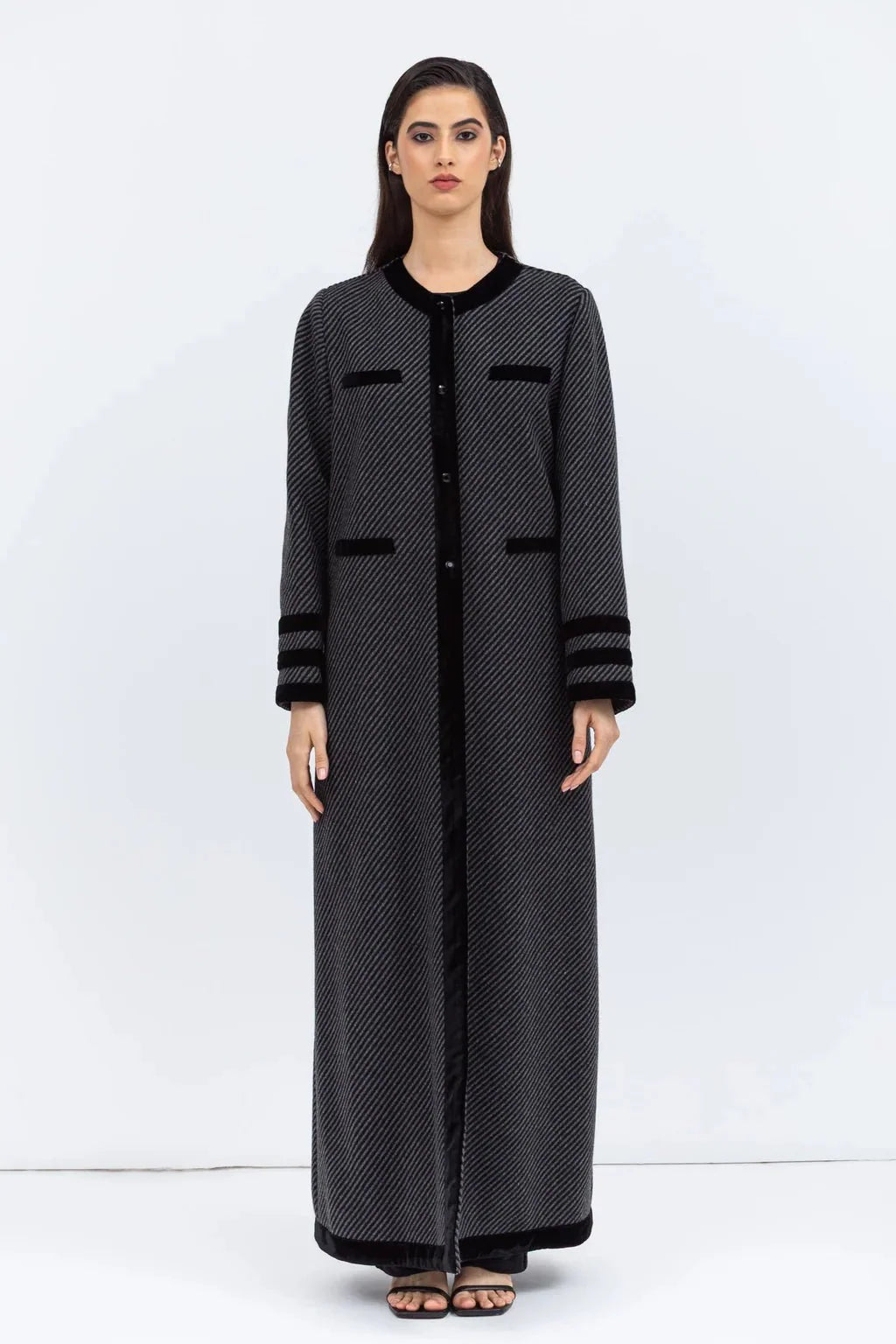 Model in a long grey wool coat abaya with black velvet trim and patch pockets, against a white wall