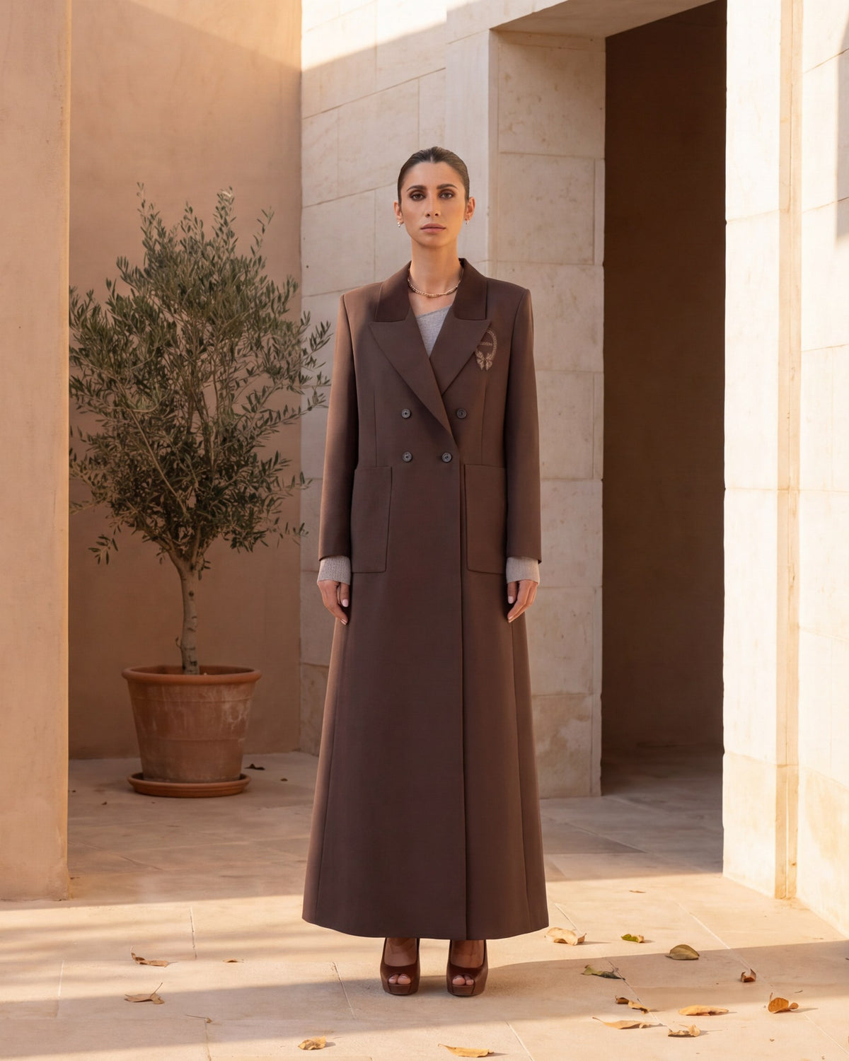 Model in a saddle brown double-breasted crepe abaya coat in a sunlit limestone courtyard
