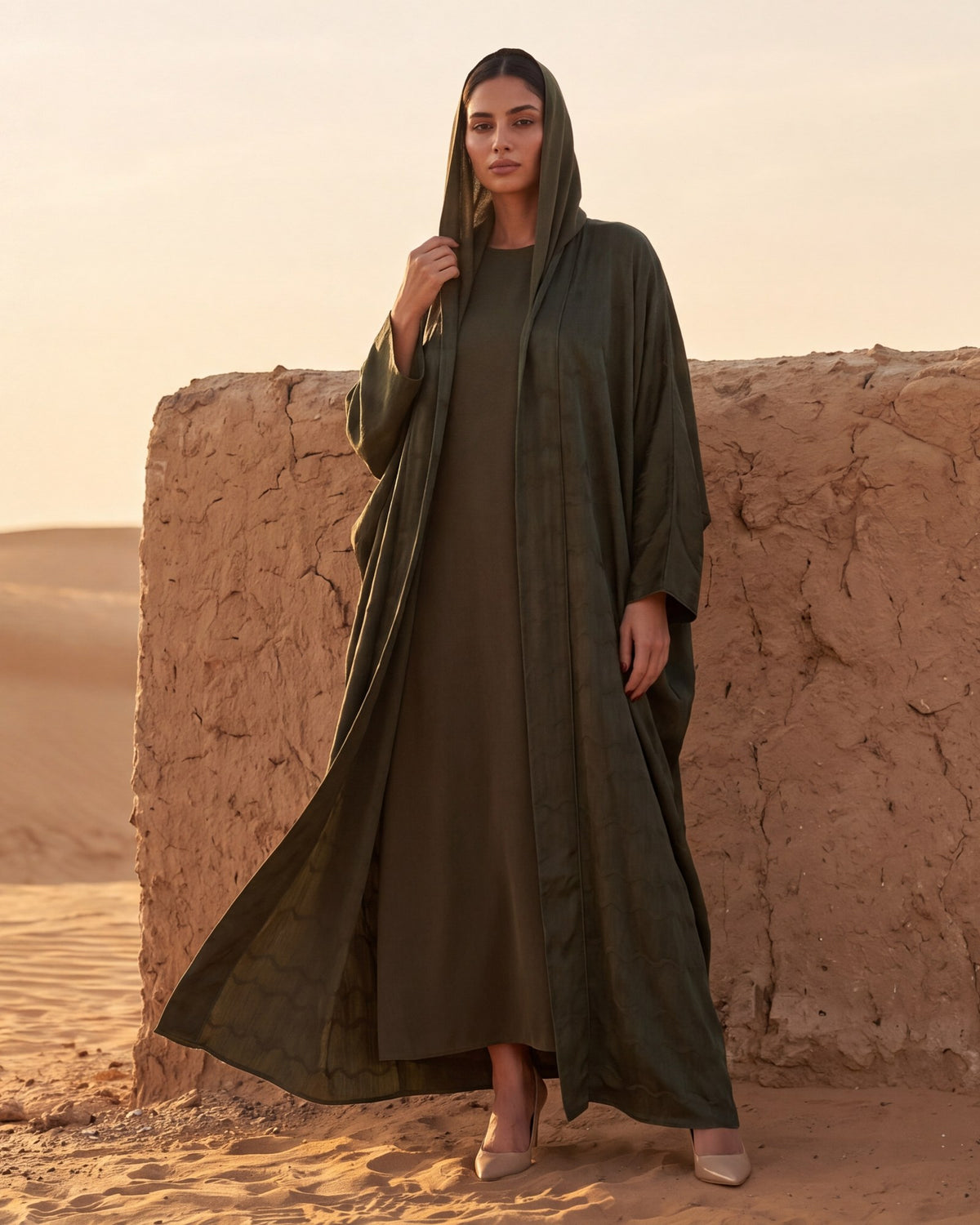 Model in a dark olive washed-silk hooded abaya beside a mud-brick wall at the edge of the desert