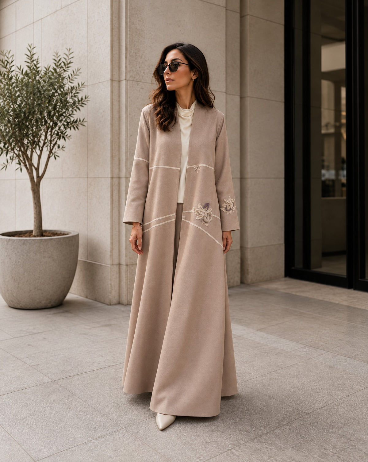 Model in a long beige silk abaya with ivory piping and appliqué flowers, outside a stone building