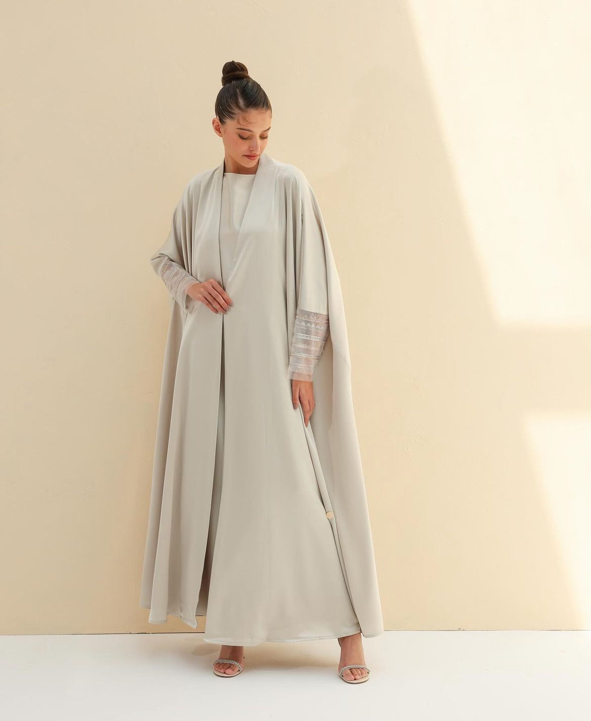 Model in a pale beige silk abaya set with sheer embellished cuffs, against a cream wall
