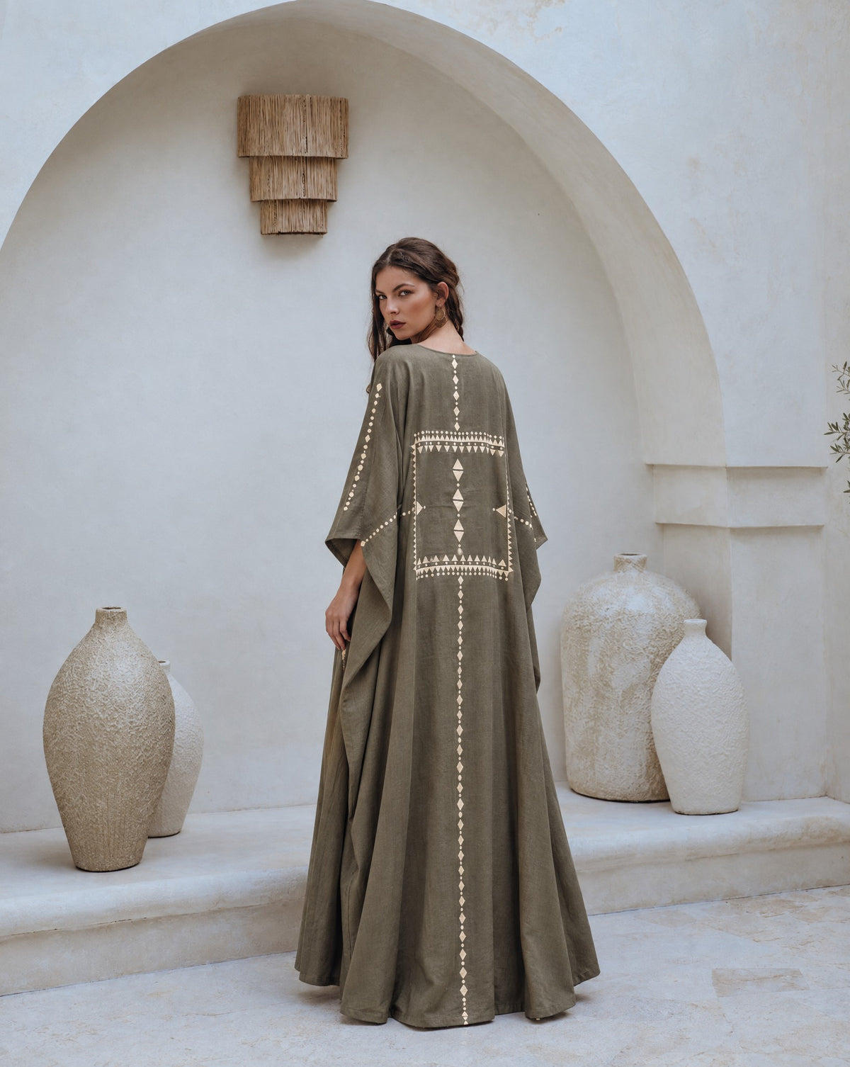 Back view of a model in a dark olive abaya set with a cream embroidered panel, in a white arched courtyard