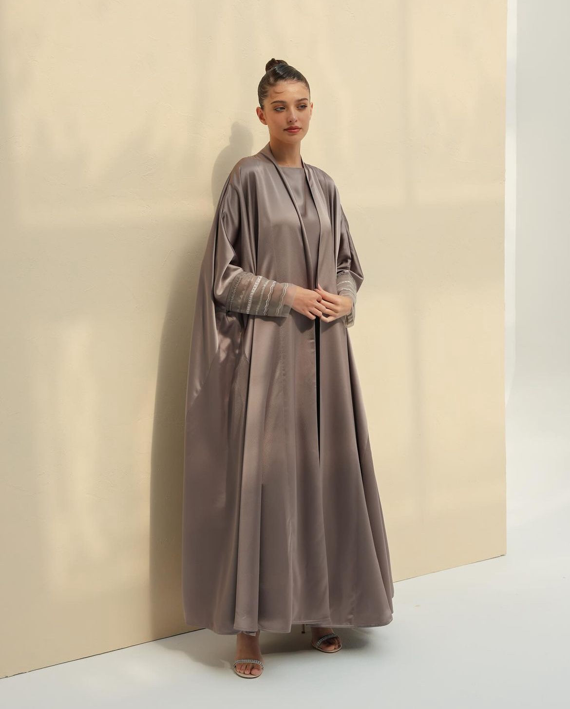 Model in a taupe silk abaya set with sheer embellished cuffs, leaning on a cream wall