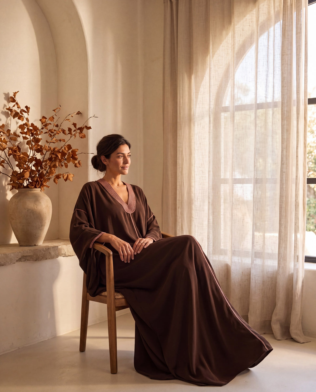 Model seated in the Naima chocolate crepe kaftan by an arched window with linen curtains