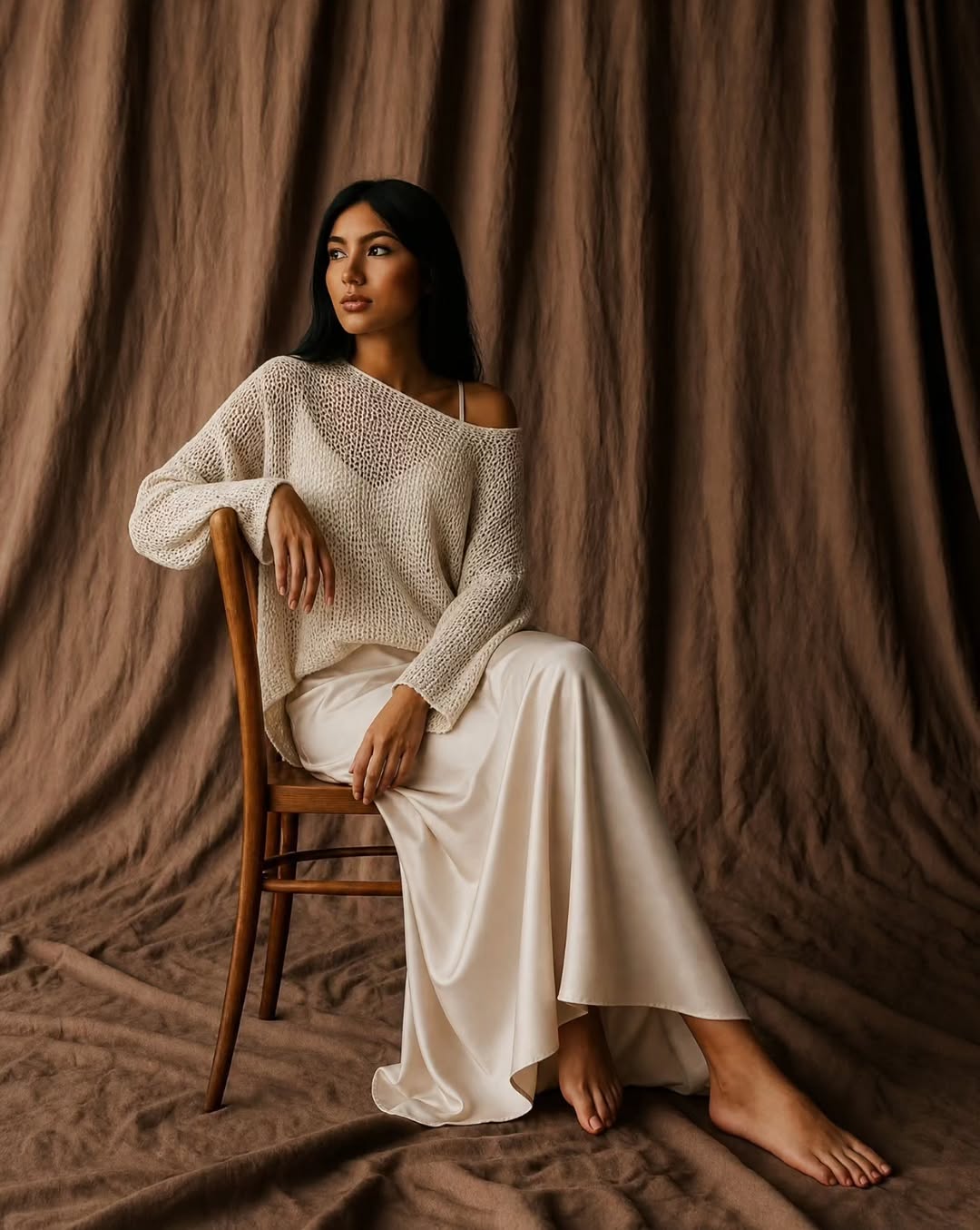 Model in a cream open-knit sweater off the shoulder and an ivory satin skirt, seated on a wooden chair against a brown drape