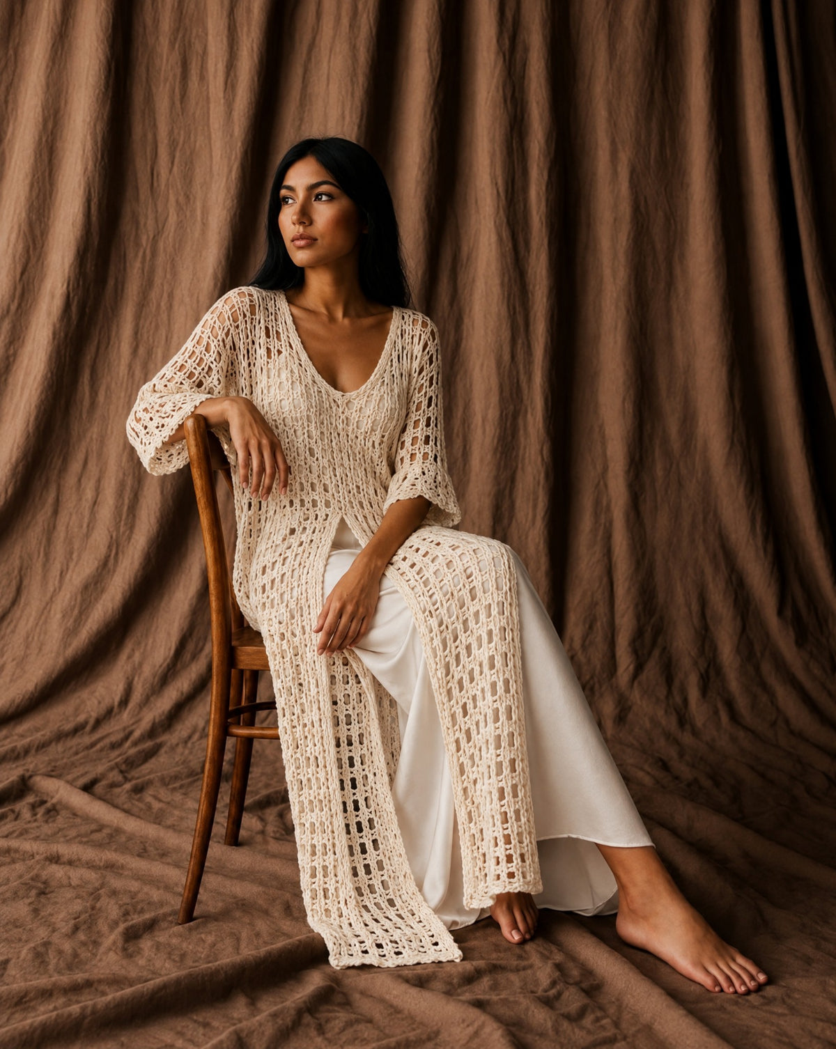 Model in a long cream crochet tunic over white trousers, seated on a wooden chair