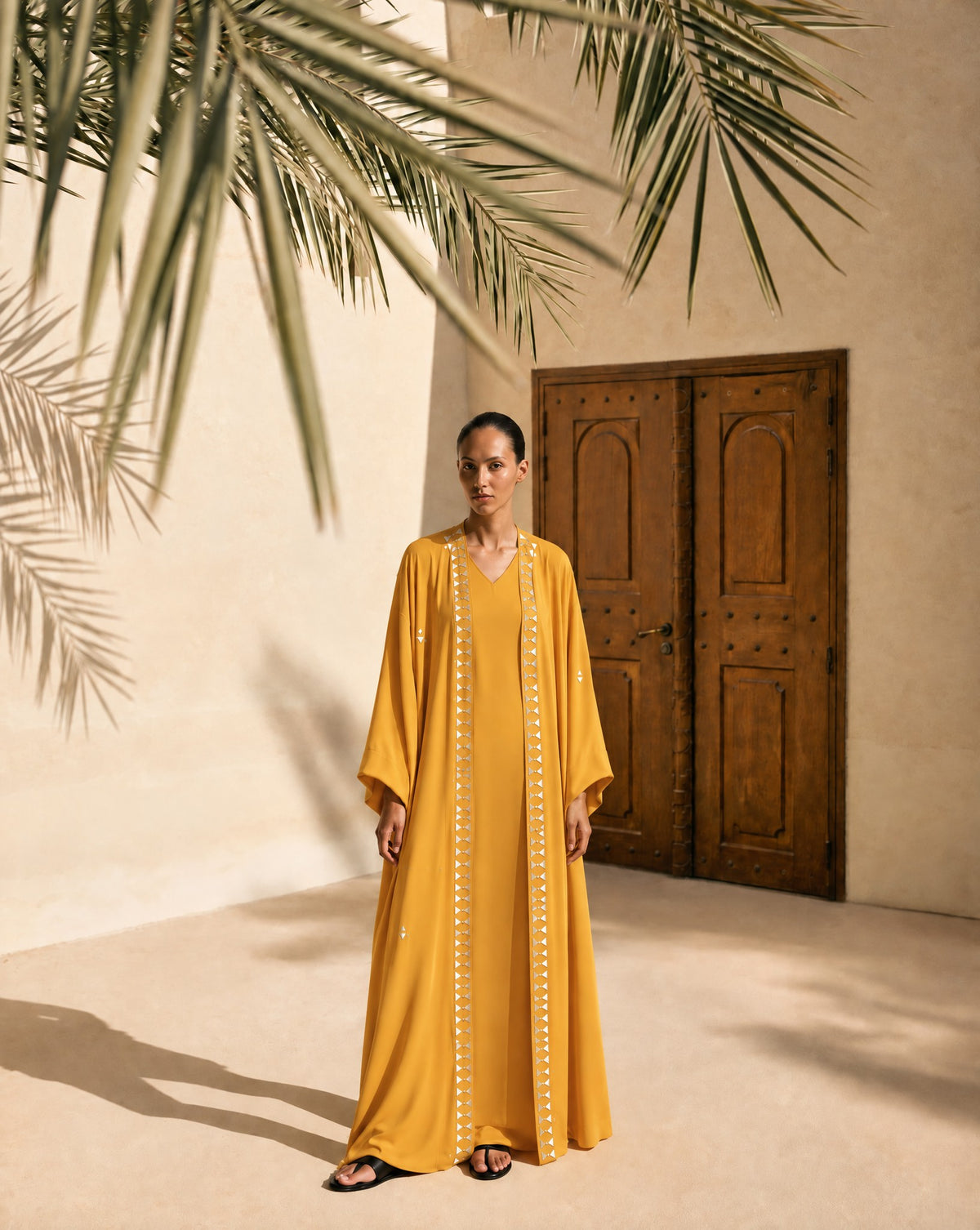 Model in a mustard georgette abaya set with white embroidered panels, under palm leaves