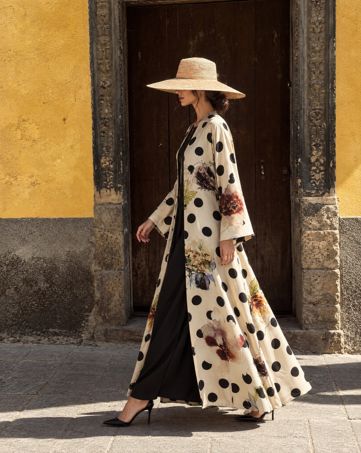 Model in a cream silk and cotton abaya with black polka dots and painted flowers, wearing a wide straw hat