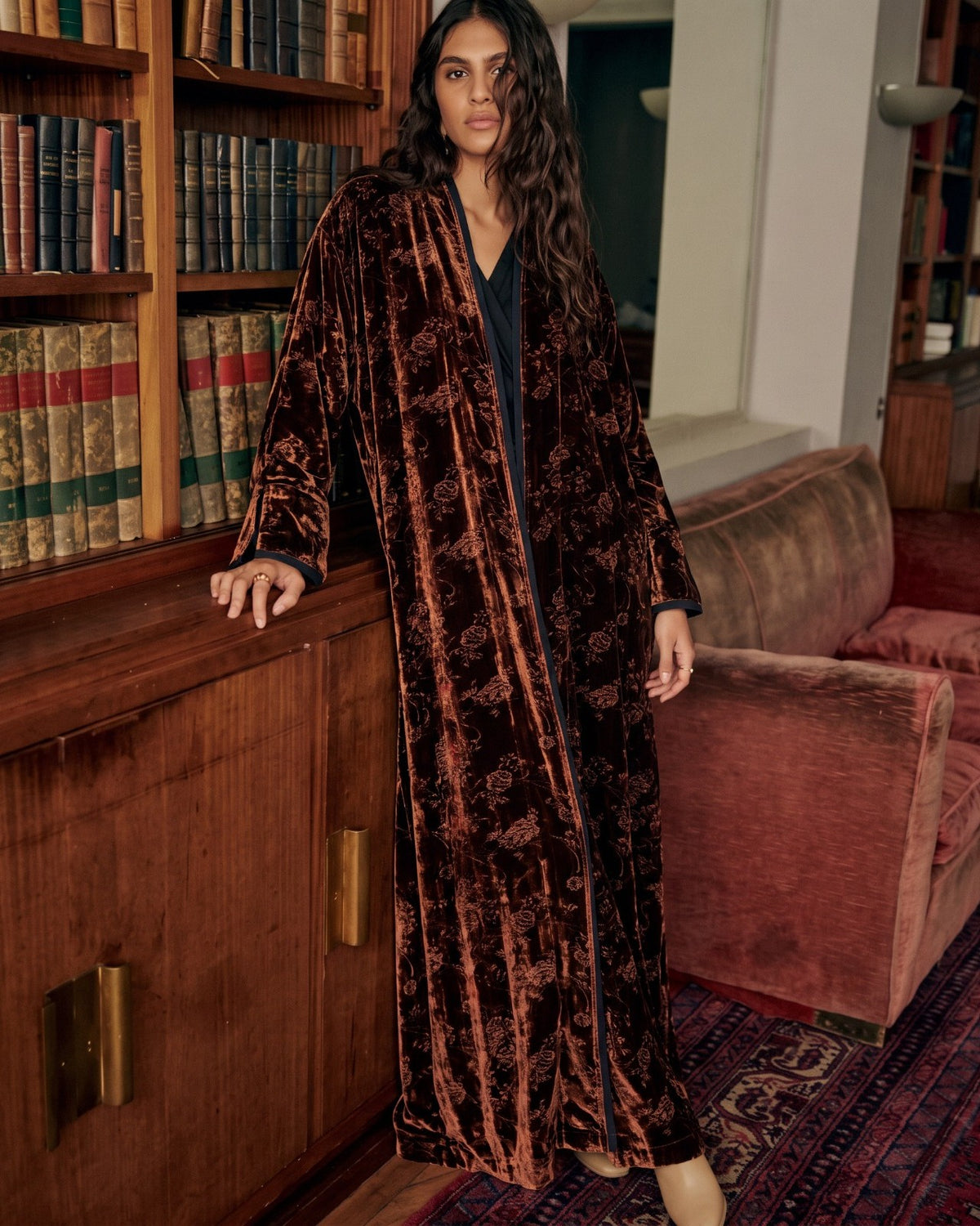 Model in an open rust floral velvet abaya over black, in a library
