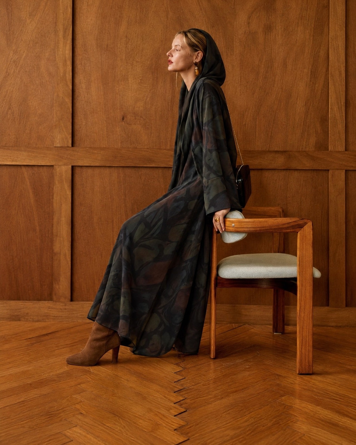 Model seated in profile in a hooded dark printed washed silk abaya, in a wood-panelled room
