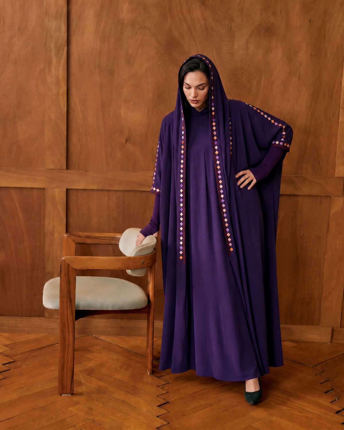 Model in a hooded purple crepe abaya with a coloured diamond trim, one hand on a wooden chair, in a wood-panelled room