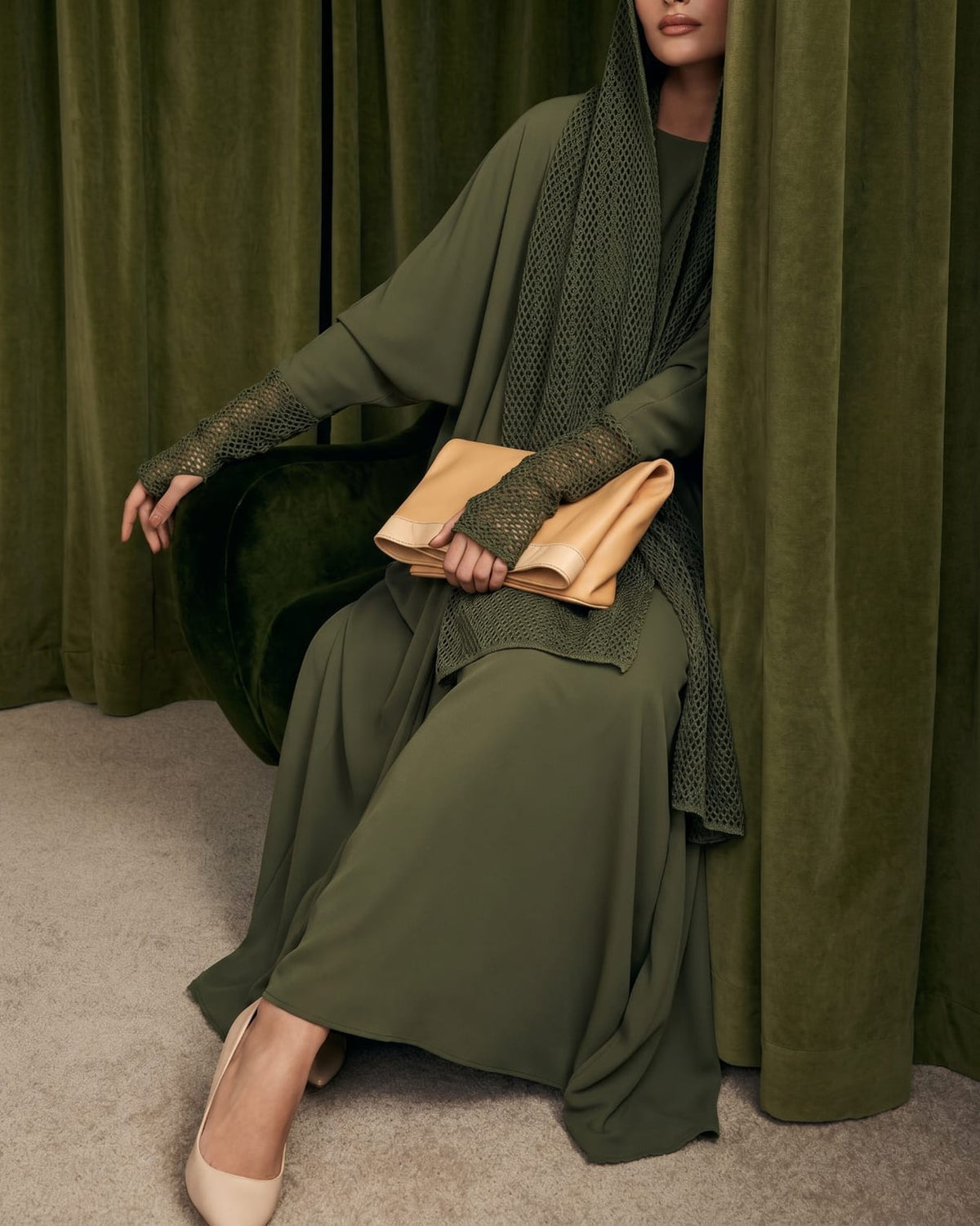 Model in an olive crepe abaya with a crochet shawl and crochet gloves, holding a tan clutch, among green curtains